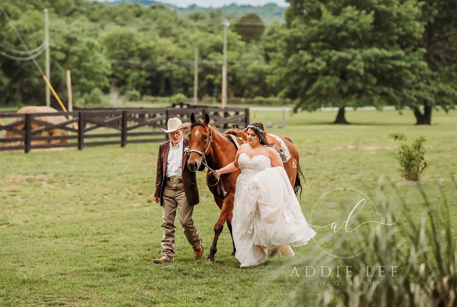 A bride in a lace gown walks a chestnut horse across a green LIKAZOO pasture, led by a handler in a cowboy hat.