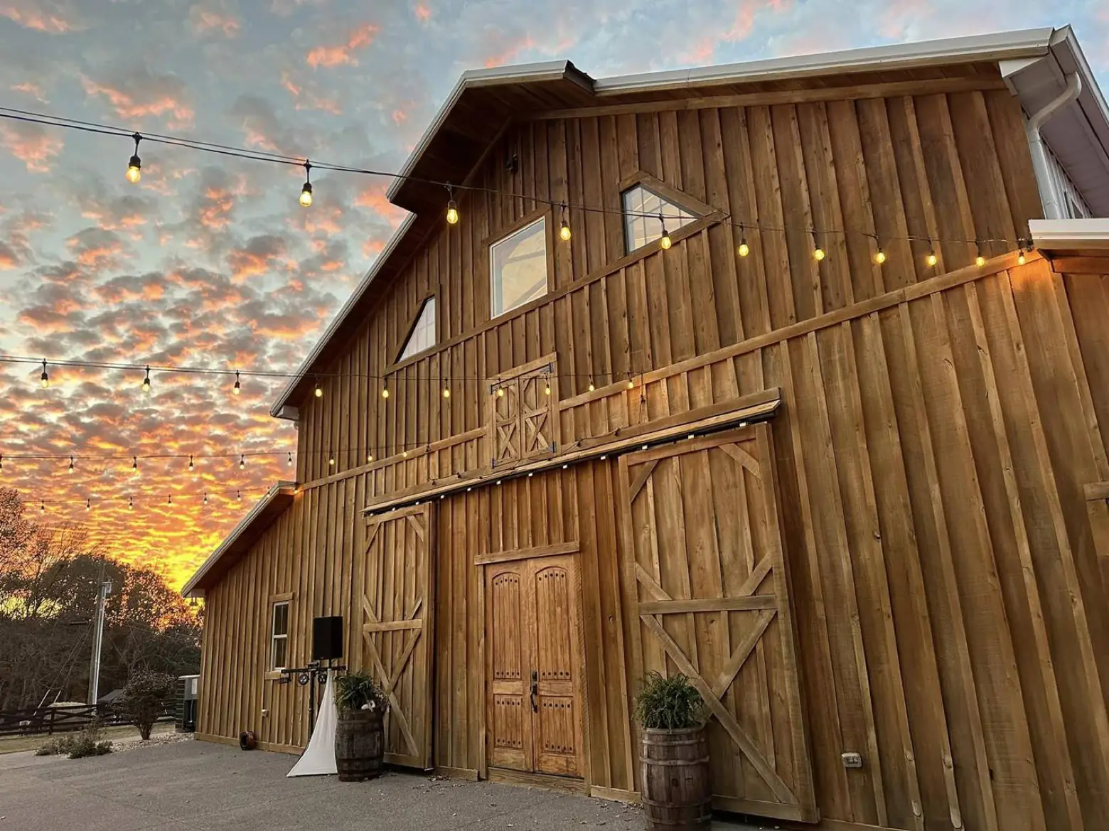 The wedding barn at LIKAZOO at golden hour — warm wood siding, sliding barn doors, and strings of café lights against a coral-and-blue Tennessee sunset.