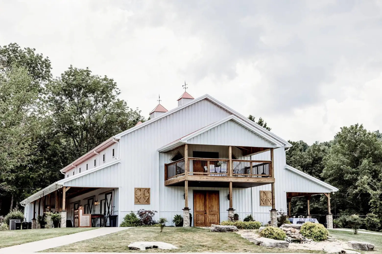 The white wedding barn at LIKAZOO — balcony, copper cupolas, and a landscaped path leading to the doors, framed by green trees.