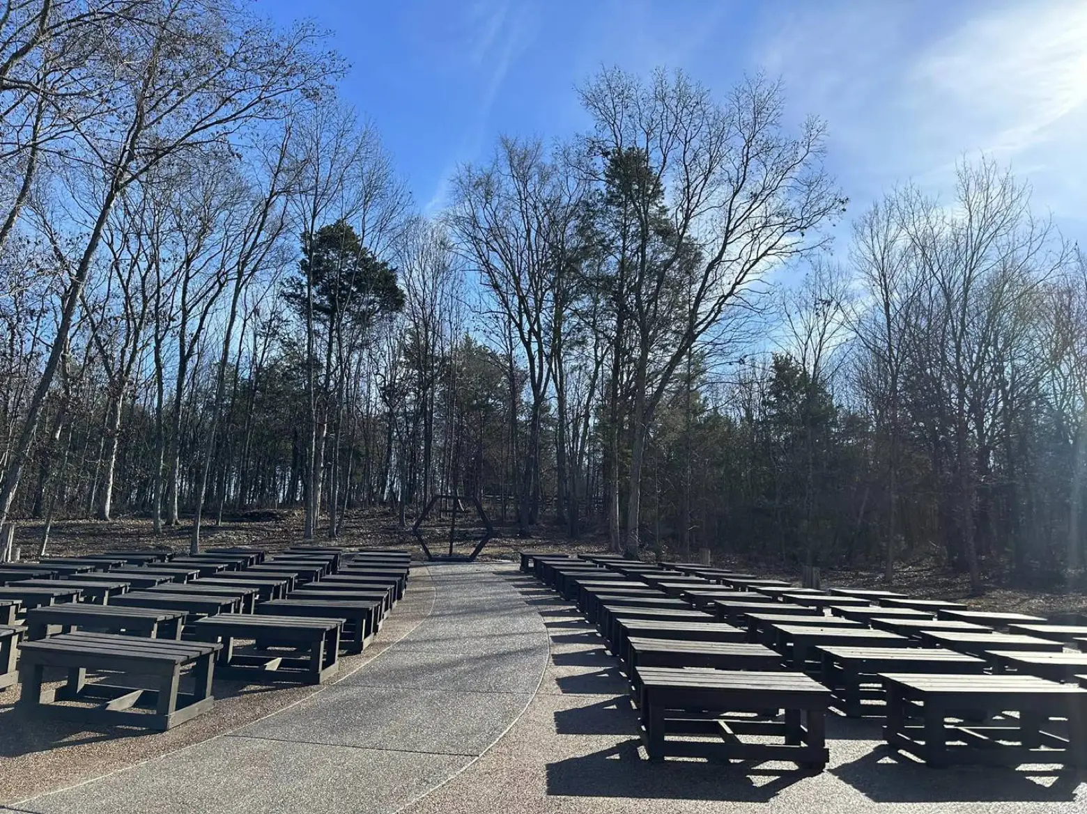 The outdoor ceremony area at LIKAZOO — rows of dark wooden benches along a curved aisle leading to a hexagonal arbor, framed by tall trees under an open sky.