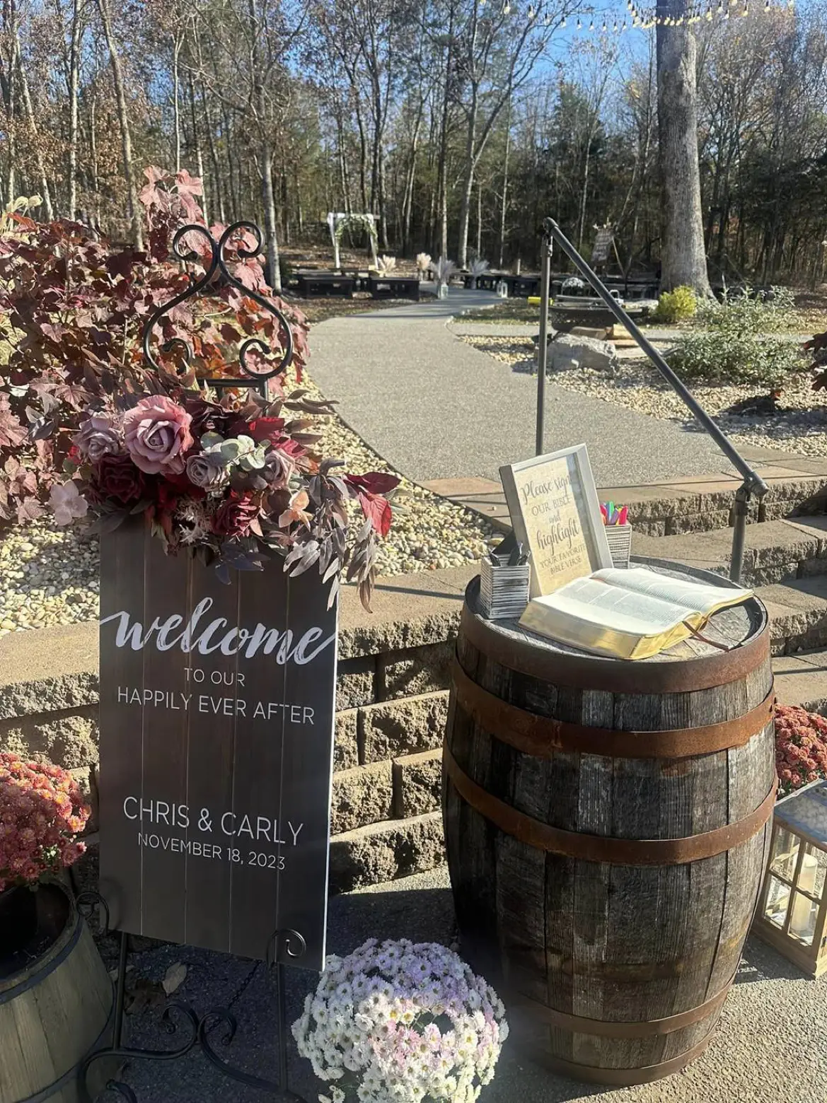 A welcome detail at a LIKAZOO wedding — a hand-lettered welcome sign beside an open Bible and pens on a whiskey barrel, the ceremony grounds beyond.