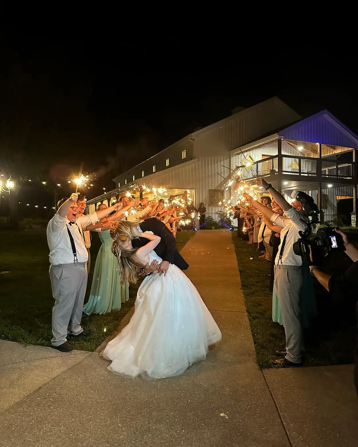 A night-time sparkler send-off outside the lit white barn at LIKAZOO — guests line a path holding sparklers as the couple shares a dip under the glowing balcony.
