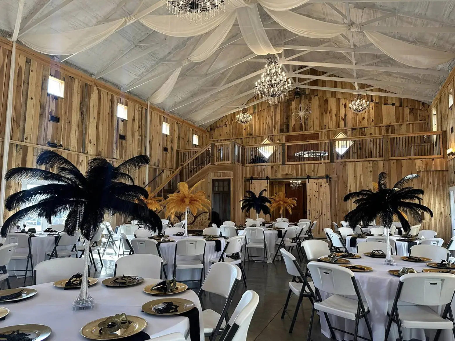A styled reception inside the barn at LIKAZOO — white-linen round tables with tall centerpieces, a wide dance floor, and a lofted staircase under chandeliers.