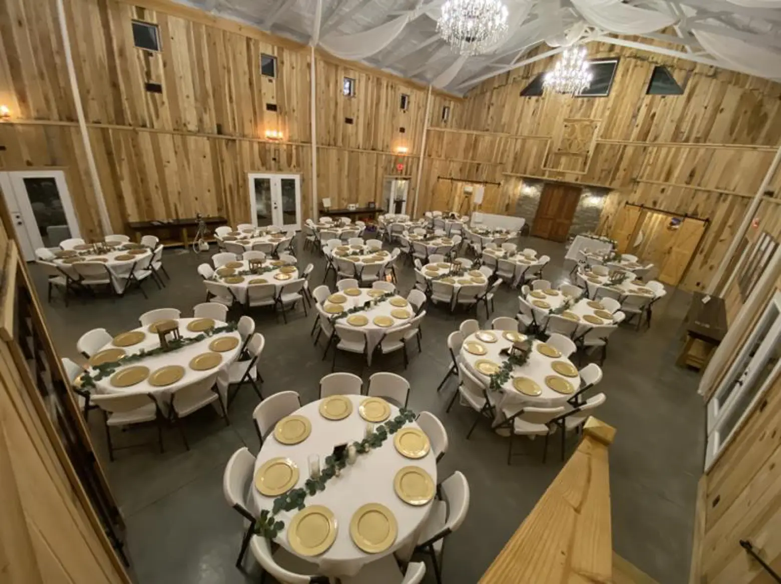 Inside the white barn at LIKAZOO — round tables set with gold chargers and greenery runners under draped ceiling fabric and crystal chandeliers, warm pine walls all around.