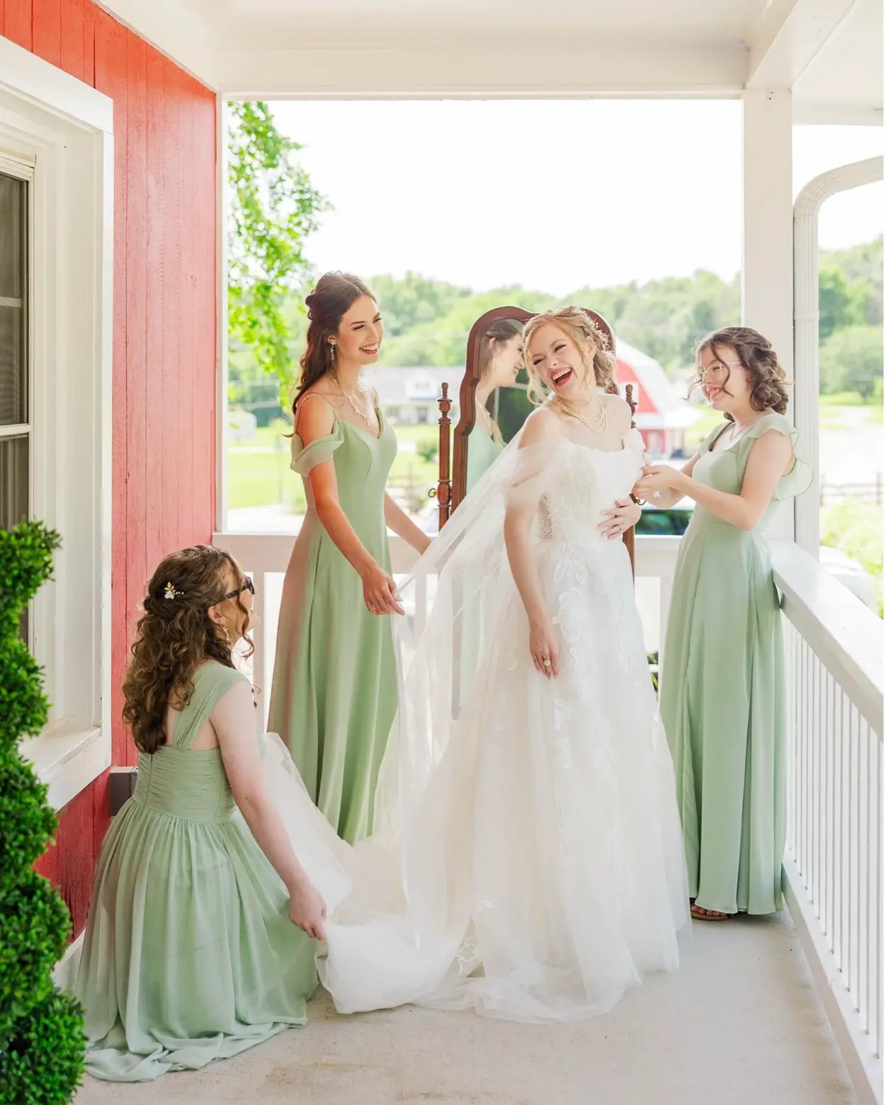 A bride laughing with her bridesmaids in sage-green dresses on a farmhouse porch at LIKAZOO, the red barn and green fields behind them.